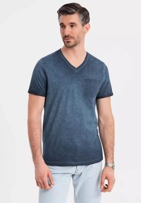 T-shirt navy a V in un tessuto sbiadito e melangiato. Maniche corte con una tasca applicata sul lato sinistro del petto. Modello che indossa jeans azzurri.