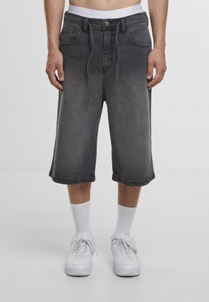 Mężczyzna ubrany w wyblakłe czarne dżinsowe spodenki do kolan, białe skarpetki i białe trampki, stojący na jednolitym szarym tle.