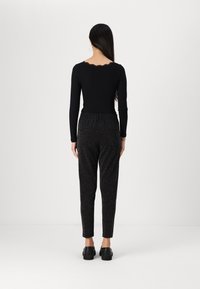 Haut à manches longues noir avec un col festonné, associé à un pantalon noir ajusté et scintillant ; modèle portant des chaussures noires. Fond simple.