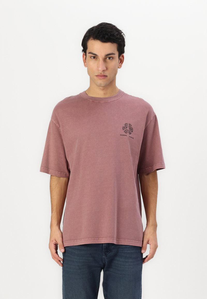 Camiseta de manga corta sencilla en malva deslavado con cuello redondo. Presenta un pequeño gráfico negro en el lado izquierdo del pecho. Dobladillo cosido.