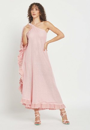 Žena v ružových maxi šatách s jedným ramenom a volánikmi na okrajoch, obutá v bielych sandáloch na vysokom opätku so remienkami, stojaca pred jednofarebným pozadím.