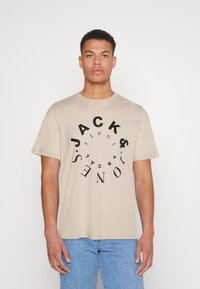 Bežs kokvilnas T-krekls ar īsām piedurknēm, ar apaļu melnu logo dizainu, kurā ir teksts "JACKS & JONES" dažādās orientācijās.