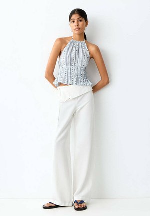 Jeune femme aux cheveux foncés attachés en queue de cheval, portant un haut sans manches à motifs avec col halter, un pantalon blanc à jambes larges et des sandales bleues, debout contre un mur blanc.