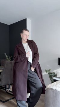Manteau long bordeaux foncé avec un col cranté, orné de boutons. Porté sur une chemise claire et un pantalon gris ample dans un environnement intérieur moderne.