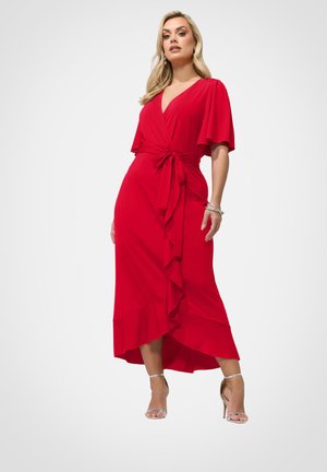 Vrouw draagt een rode wikkeljurk met vleermuismouwen, taillekoord, geruffleerde hoge-lage zoom en zilveren sandalen met hoge hakken, staand tegen een witte achtergrond.