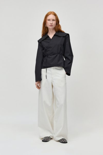 Femme aux cheveux rouges portant un haut noir à manches longues avec un cordon à la taille, un pantalon blanc ample et des chaussures noires cloutées, debout devant un fond uni.