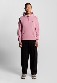 Felpa rosa con tasca frontale e cappuccio regolabile, con un piccolo logo a forma di farfalla gialla. Abbinata a pantaloni neri larghi e sneakers.