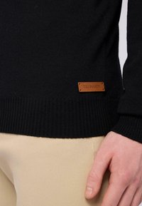 Černý pletený svetr s ribovanými manžetami a lemem, se malou hnědou koženou etiketou s nápisem "TRUSSARDI" po straně. Nosený s béžovými kalhotami.