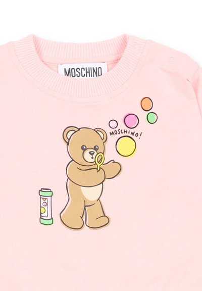 Różowy strój z misiem wydmuchującym kolorowe bańki mydlane w pobliżu pojemnika na bańki, z napisem "MOSCHINO" nad misiem.