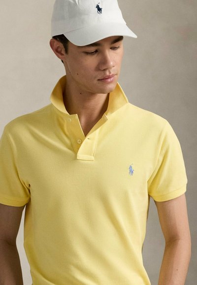 Polo giallo con colletto classico, caratterizzato da un discreto logo ricamato. Indossato con un cappellino bianco che mostra un logo simile.
