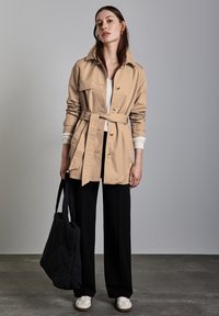 Beige trench coat, johon vyö on lisätty, isoilla napeilla, käännetyillä hihoilla ja pehmeällä tekstuurilla, yhdistettynä mustiin leveälahkeisiin housuihin ja mustaan laukkuun.