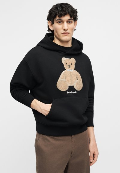 Jonge man die een zwarte hoodie draagt met een beige teddybeervormig ontwerp en de tekst "Palm Angels", bruine broek, staand met hand in zak.