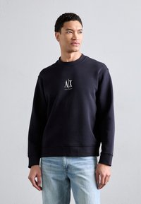 Felpa blu navy con collo a giro, caratterizzata da un logo bianco al centro. Tessuto morbido, polsini a costine e orlo dritto. Abbinata a jeans blu chiaro.