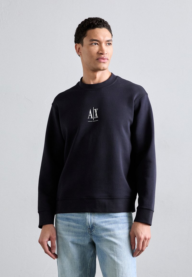 Felpa blu navy con collo a giro, caratterizzata da un logo bianco al centro. Tessuto morbido, polsini a costine e orlo dritto. Abbinata a jeans blu chiaro.