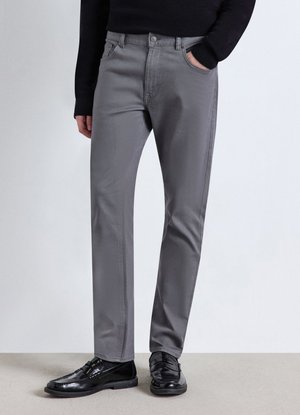 Ein Mann trägt graue gerade geschnittene Hosen, einen schwarzen Pullover und schwarze Lederloafers und steht mit einer Hand in der Tasche.