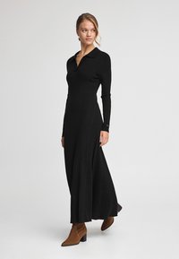 Schwarzes, geripptes Kleid mit Kragen, langen Ärmeln und einem schwingenden Rock. Getragen mit braunen Stiefeletten, die eine strukturierte Oberfläche haben.