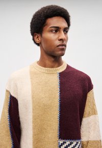 Kootud kampsun, millel on mitmevärvilised paneelid beežis, pruunis ja burgundias, soonikkoeline struktuur, palmikutega detailid ning sinine õmblusdetail.