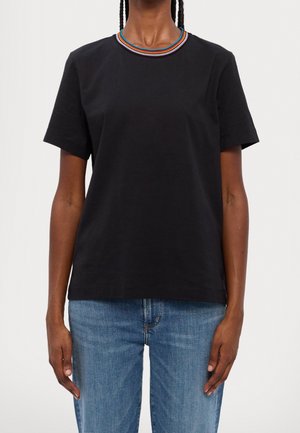 Persona vistiendo una camiseta negra de manga corta con un cuello a rayas multicolor combinada con vaqueros azules, de pie frente a un fondo liso.