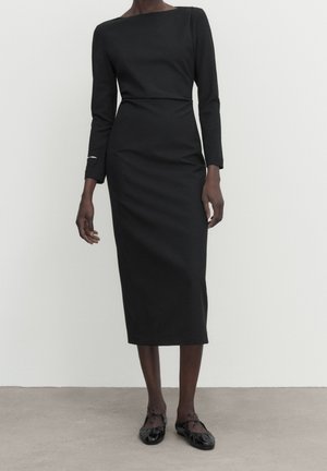 Woman wearing a fitted, long-sleeve black midi dress with boat neckline and black ballet flats, standing against a plain background.