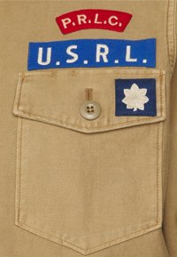 Camisa de tela beige con un bolsillo en el pecho que tiene un botón beige. Tres parches bordados: "P.R.L.C." en rojo, "U.S.R.L." en azul, y un cuadrado azul con una hoja blanca.