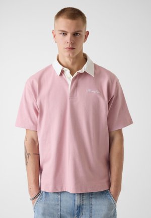 Giovane con capelli corti che indossa una polo rosa chiaro con colletto bianco e jeans azzurri, in piedi davanti a uno sfondo semplice.