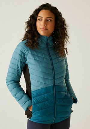 Frau mit lockigem Haar, die eine gesteppte Jacke in Türkis und Dunkelblau trägt, die Hände in den Taschen hat und leicht seitlich blickt, vor einem schlichten Hintergrund.