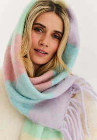 Kobieta o blond włosach nosząca pastelowy, paskowany kaptur z szalikiem i kremowy sweter, patrząca łagodnie w stronę kamery.