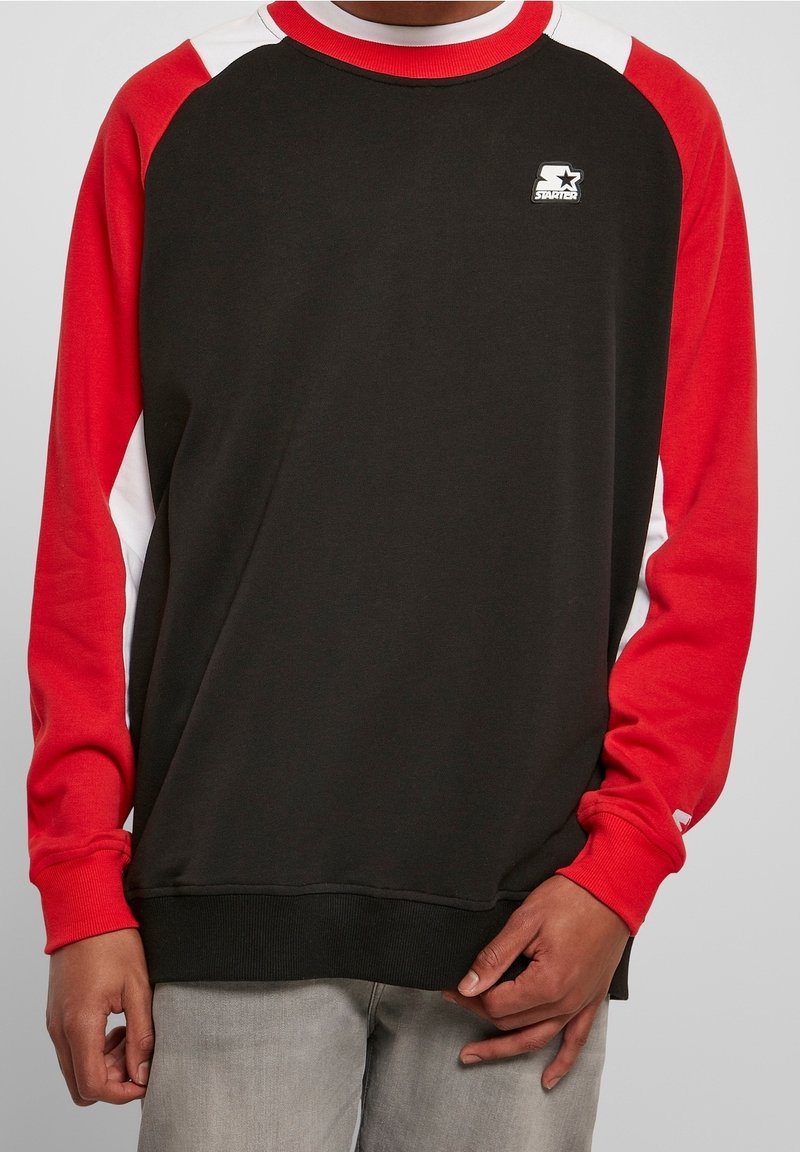 Sudadera negra con mangas rojas y detalles en blanco, con un pequeño logo blanco de Starter en el pecho, usada con pantalones gris claro.