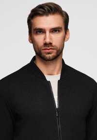 Czarna bomberka z zamkiem z przodu i zaokrąglonym kołnierzem. Materiał ma gładką fakturę. Nosi się ją na jasnej koszuli.