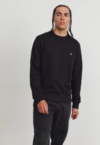 Juodas apvalios iškirptės džemperis su struktūruota apdaila, turintis mažą logotipą ant krūtinės. Pora su tamsiomis džinsomis, minimalistiškas dizainas.