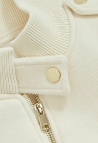 Valkoinen fleece-takki, jossa on ribattua kaulusta, kultaisia neppareita ja hopeisia vetoketjuja. Pehmeä rakenne ja siisti muotoilu.