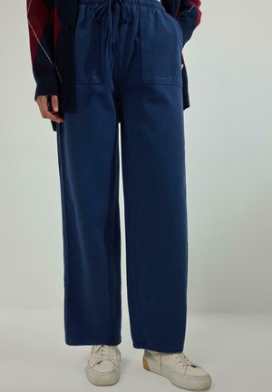 Persona che indossa pantaloni larghi blu navy con coulisse, tasche frontali e sneakers bianche, in piedi davanti a uno sfondo neutro.