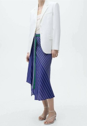Persoon die een witte blazer, beige top, blauw-wit gestreepte rok met groene bies en beige riempjessandalen met hak draagt, staand op een witte achtergrond.