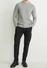 Pull tricot gris avec un col rond et des poignets côtelés, associé à un pantalon cargo noir doté de poches latérales et d'une texture robuste. Des chaussures noires complètent la tenue.