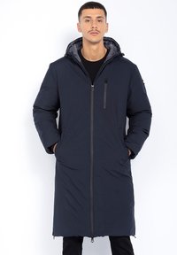 Mann trägt einen langen, dunkelblauen, kapuzenlosen Wintermantel mit Reißverschluss-Taschen und steht mit Händen in den Taschen vor einem schlichten weißen Hintergrund.