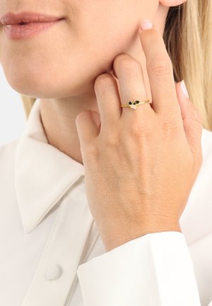 Anillo de oro con gemas verdes y azules, diseño delicado, llevado en una mano con un acabado natural, combinado con una camisa blanca con cuello.