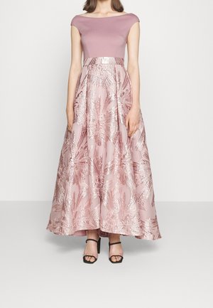 Kvinde iført en lyserød off-shoulder kjole med metalliske blomsterprint og sorte åbne høje hæle sandaler, stående mod en ensfarvet baggrund.