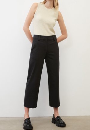 Femme portant un haut sans manches crème, un pantalon large noir coupé au dessus de la cheville et des mocassins noirs épais, debout contre un mur blanc.