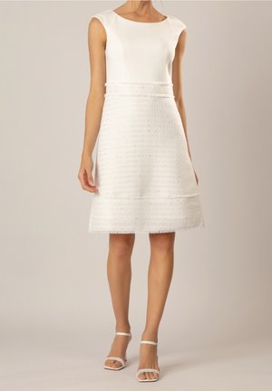 Kobieta w białej sukience bez rękawów z teksturowaną spódnicą w kształcie litery A oraz białych butach na obcasie z paskiem wokół kostki, stojąca na tle jednolitego beżowego tła.