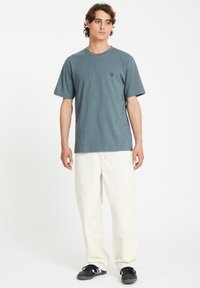 T-shirt gris-bleu à manches courtes avec un col rond et un petit logo noir, accompagné d'un pantalon ample crème et de baskets noires.