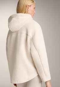 Giacca con cappuccio in pile morbido color crema, dal taglio rilassato, con orlo arrotondato e superficie testurizzata, cuciture visibili e un design accogliente.