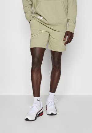 Olive green cotton sweatshirt and matching shorts, featuring a logo accent on the shorts, paired with white sneakers and socks.