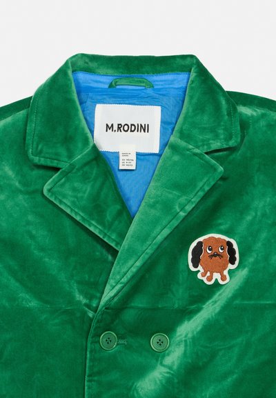 Chaqueta de terciopelo verde cruzada con forro azul, etiqueta M. Rodini y un pequeño parche de un perro marrón de dibujos animados en el pecho.