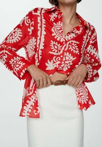 Vrouw in een rood gestreept overhemd met knopen, met een bruine riem die een witte rok ter hoogte van de taille vastmaakt.