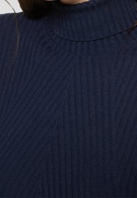 Gros plan d'une personne portant un pull en maille côtelée bleu marine avec un col montant et des motifs texturés diagonaux et verticaux.