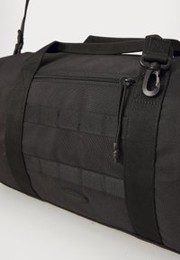 Sac de sport noir en tissu robuste, avec une poche zippée, des accents en sangle et une bandoulière ajustable. Forme rectangulaire avec un fond plat.