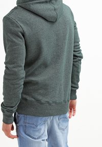 Persona vistiendo una sudadera con capucha de color verde oscuro y jeans de color azul claro, vista desde atrás contra un fondo blanco.