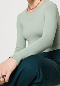 Person iført en salviegrøn langærmet trøje, mørkegrønne bukser, en kraftig sølvkæde-nikkers og en bred sølvring på venstre hånd.