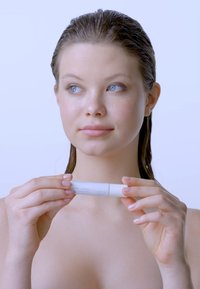 Biały kosmetyk do pielęgnacji skóry trzymany w obu rękach. Gładki cylindryczny kształt, matowe wykończenie. Model ma mokre włosy, naturalną cerę z minimalnym makijażem.