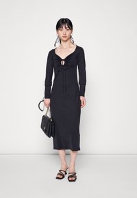 Vestido preto de mangas longas com decote em forma de coração e detalhe de cordão na frente. A modelo segura uma bolsa preta e usa sandálias pretas com tiras.